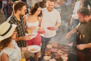 Group of friends with food surrounding a grill at summer BBQ